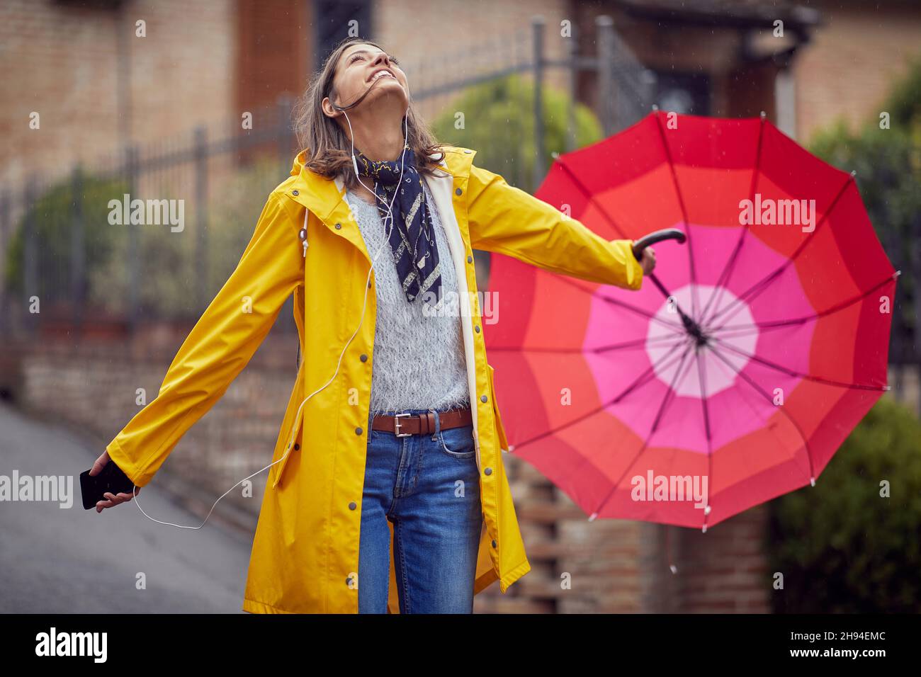 A young cheerful woman in a yellow raincoat is looking to the sky while listening to the music and walking the city in a good mood on a rainy day. Wal Stock Photo