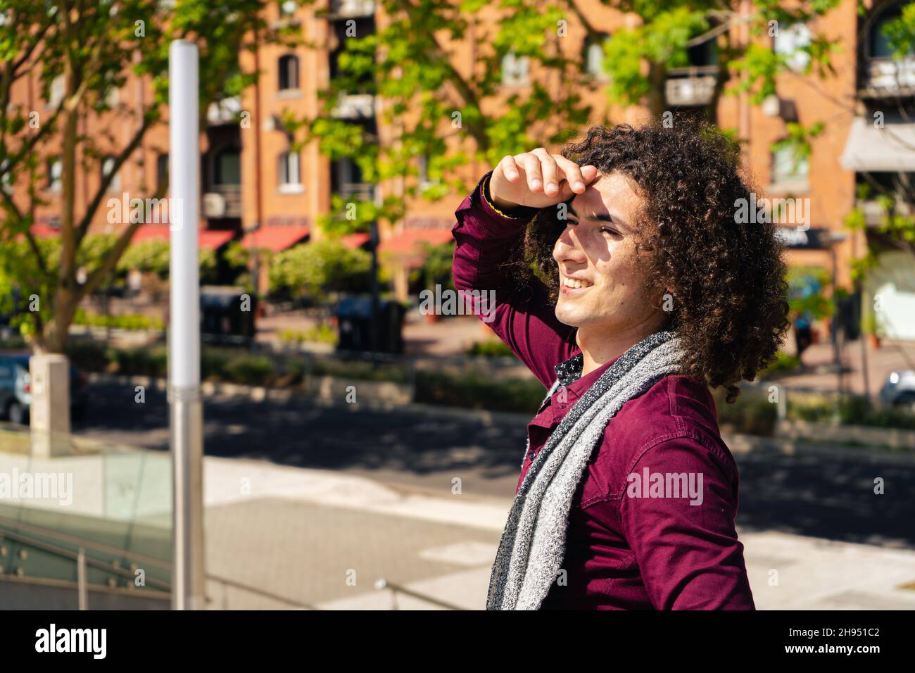 A portrait of a smiling young curly haired latin man covering his eyes from the sun and looking to his side with old buildings in the background. copy Stock Photo