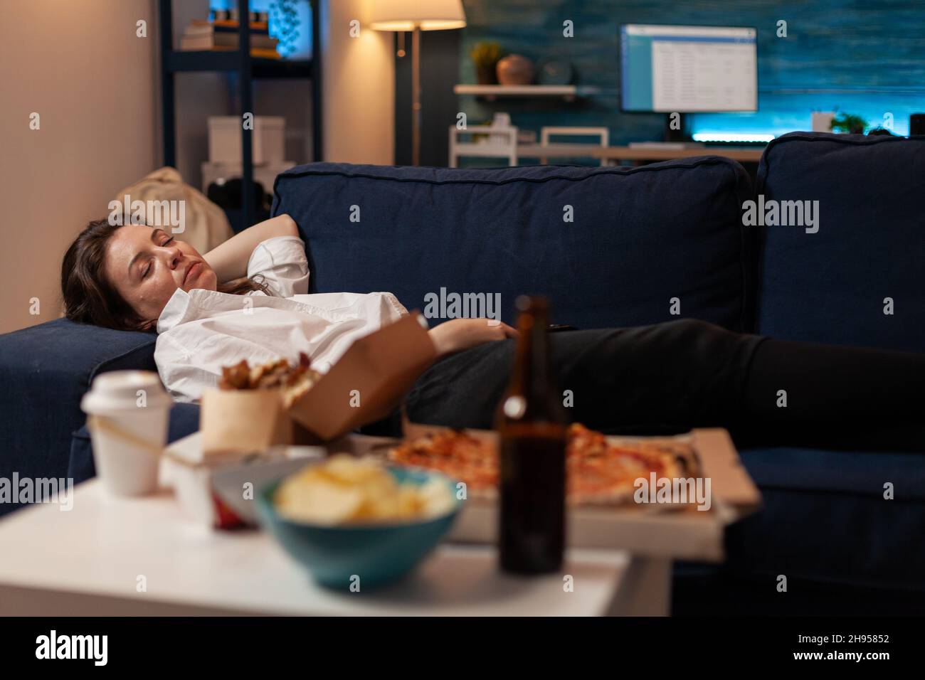 Person sleeping on sofa after drinking beer and eating home delivery pizza in front of television in living room. Tired woman falling asleep couch after large fast food takeout meal. Stock Photo