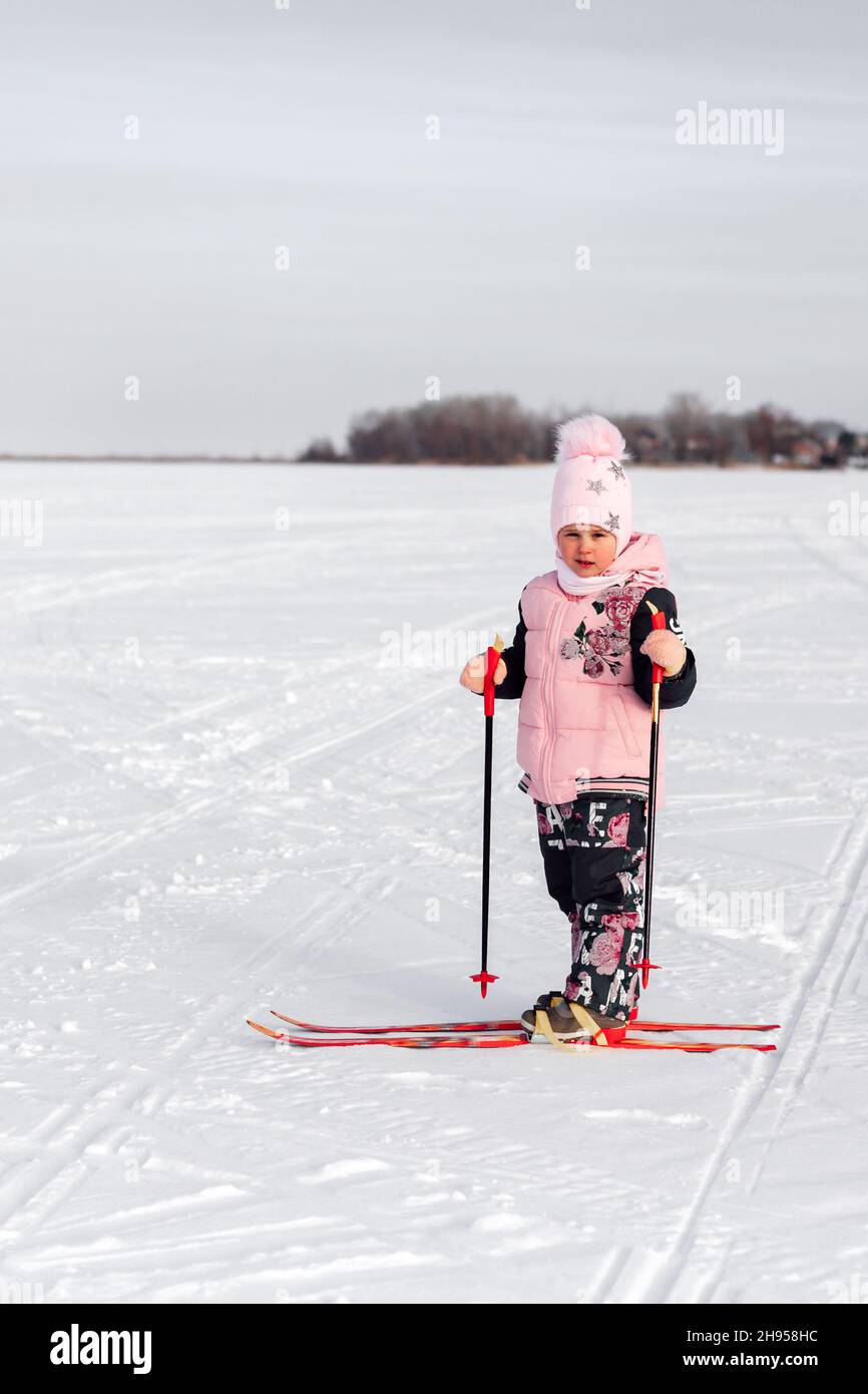 Child learns to ski. Little girl in pink suit skiing on frosty day on background of winter forest, side view, snow background Stock Photo