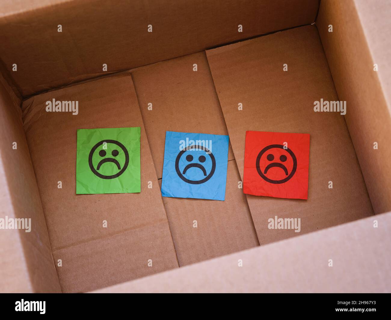 Three paper notes with sad faces on them in a cardboard box. Close up. Stock Photo