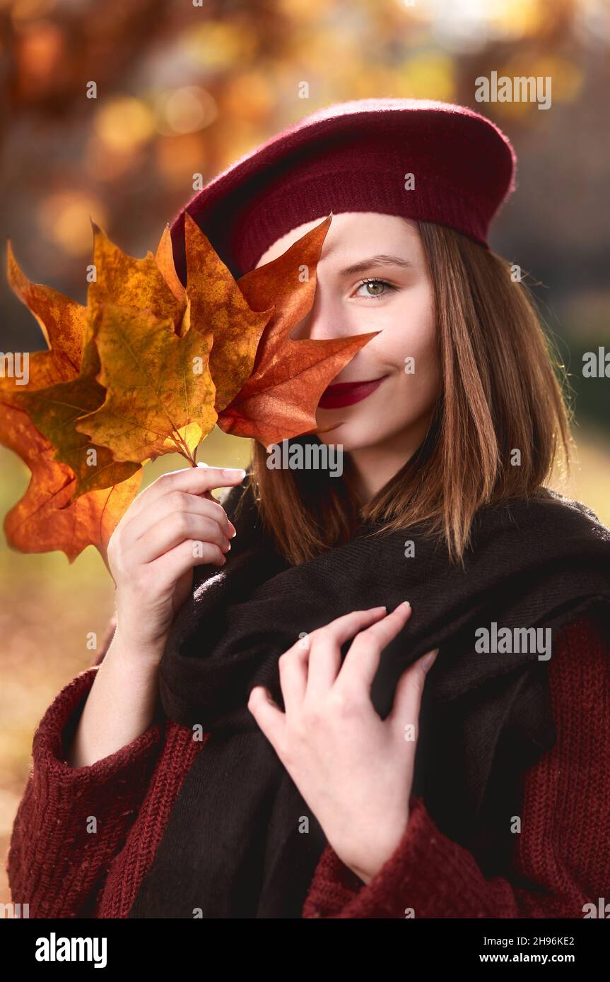 Portrait of cute smiling girl holding autumn leaves in the nature. She wears a red beret. Yellow trees and leaves at fall season in the background Stock Photo