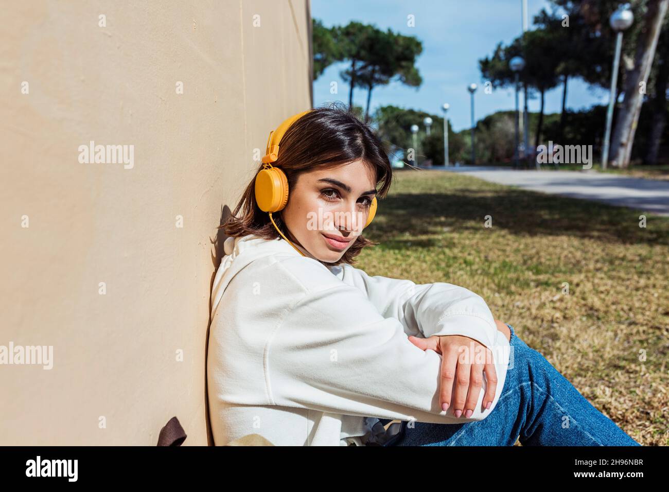 Young millennial woman listening music with headphones and looking at camera while sitting on a public park - Millennial people concept Stock Photo