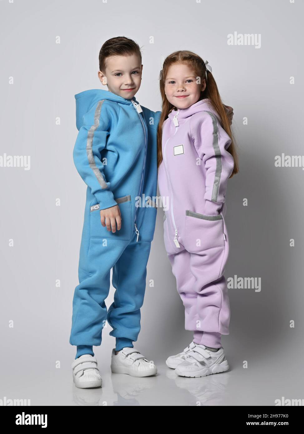 Tow smiling kids boy and girl, friends in blue and pink jumpsuits with hoods and pockets stand close together Stock Photo