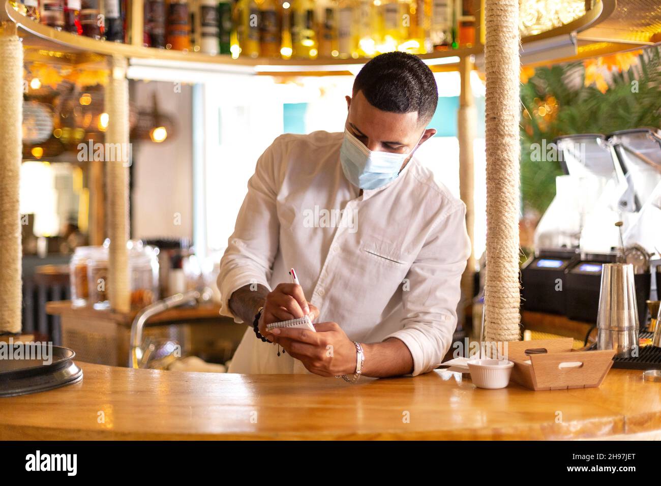 Young Latin American waiter with medical mask taking notes in his notebook at the bar of an elegant restaurant. Space for text. Stock Photo