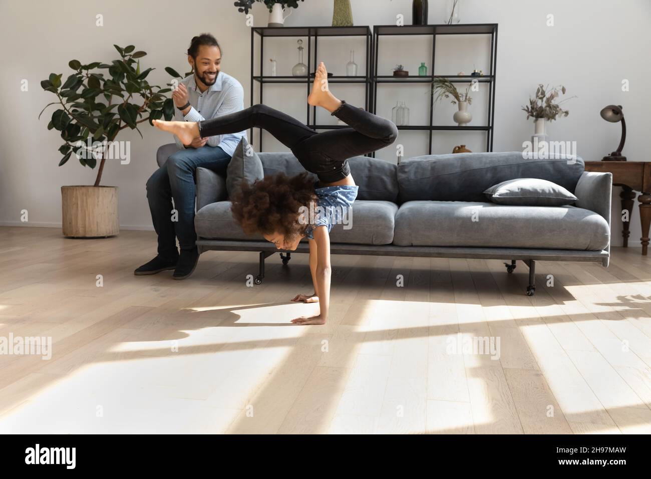 Happy young African American father watching little daughter doing acrobatics. Stock Photo