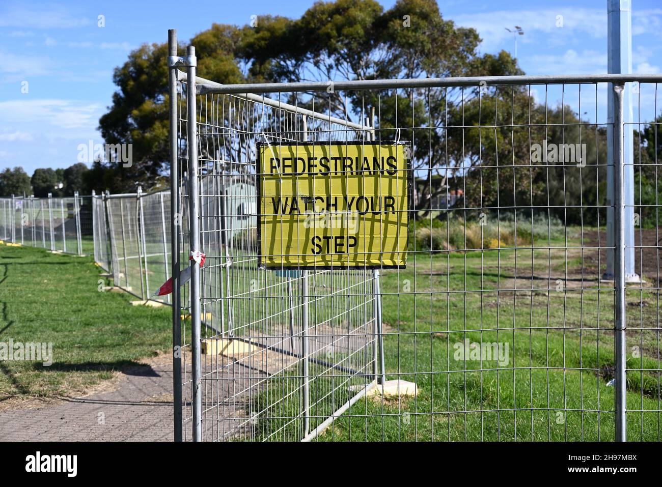 A yellow pedestrians watch your step sign, with black text, attached to a metal fence in a park Stock Photo