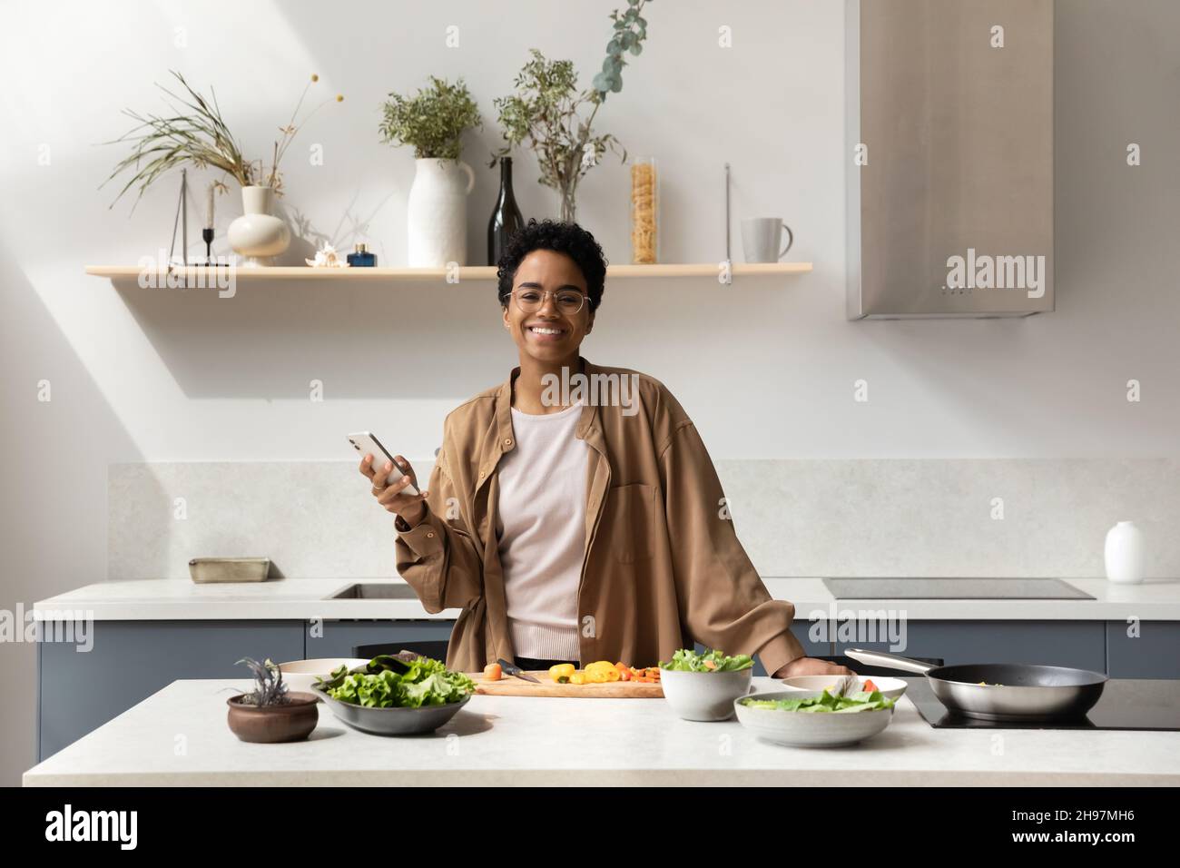 Happy African American woman holding cellphone, posing in kitchen. Stock Photo