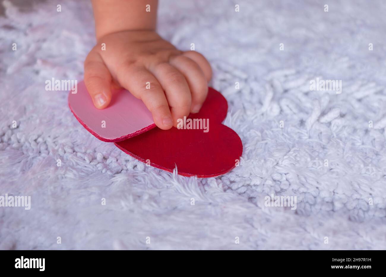 Baby's hand holds hearts on a white background. A child tries to grab hearts from a white fluffy carpet Stock Photo