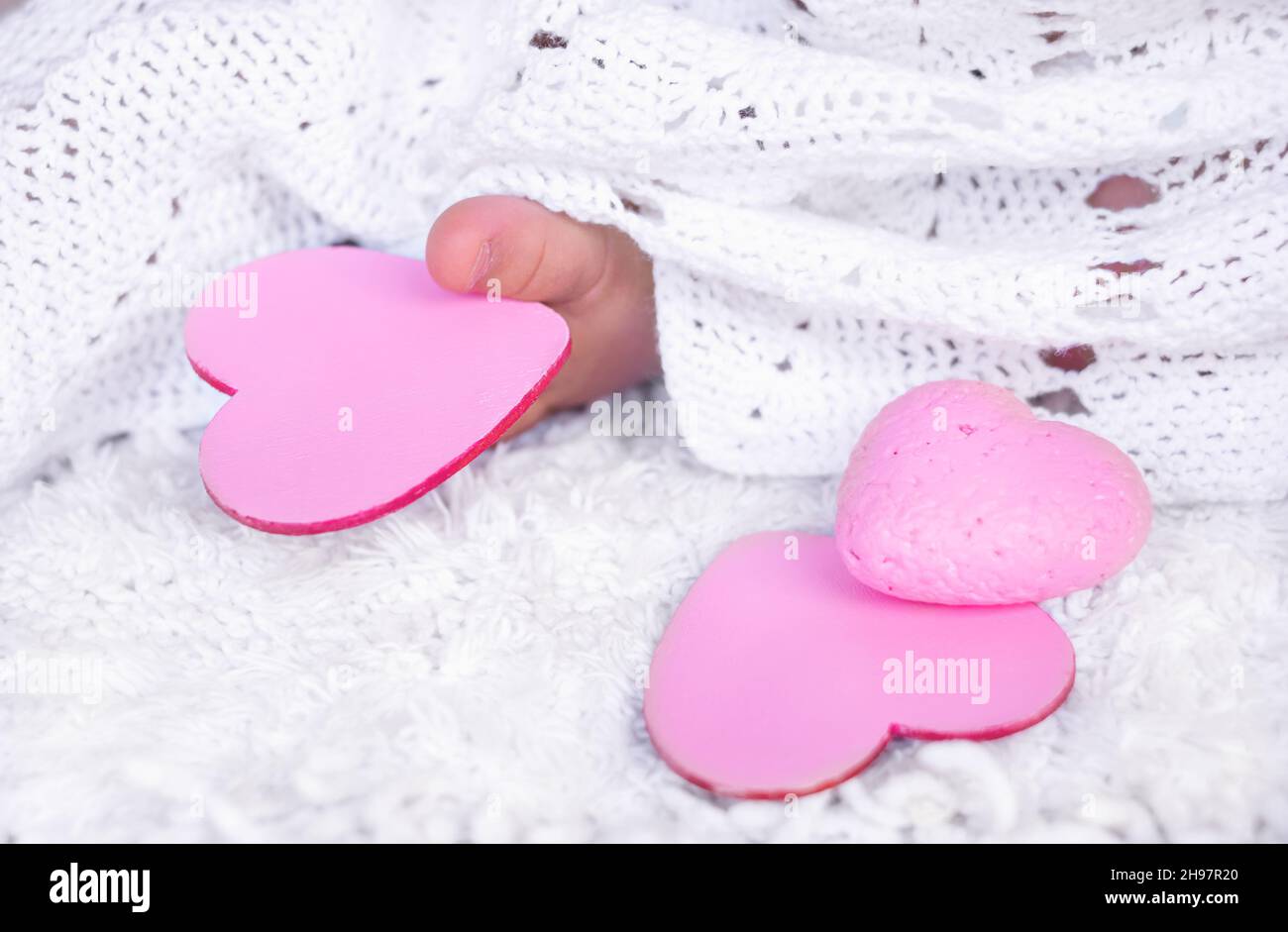 the baby's fingers are holding a pink heart. a child in a white dress holds a wooden heart with his foot Stock Photo