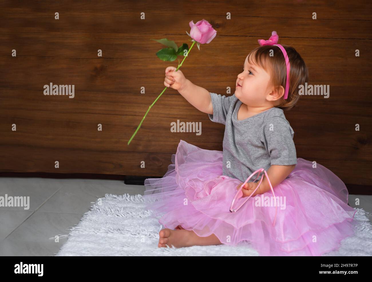 Cute baby with flower on a wooden background. Child on the floor in a pink dress, a girl looks at a pink rose Stock Photo