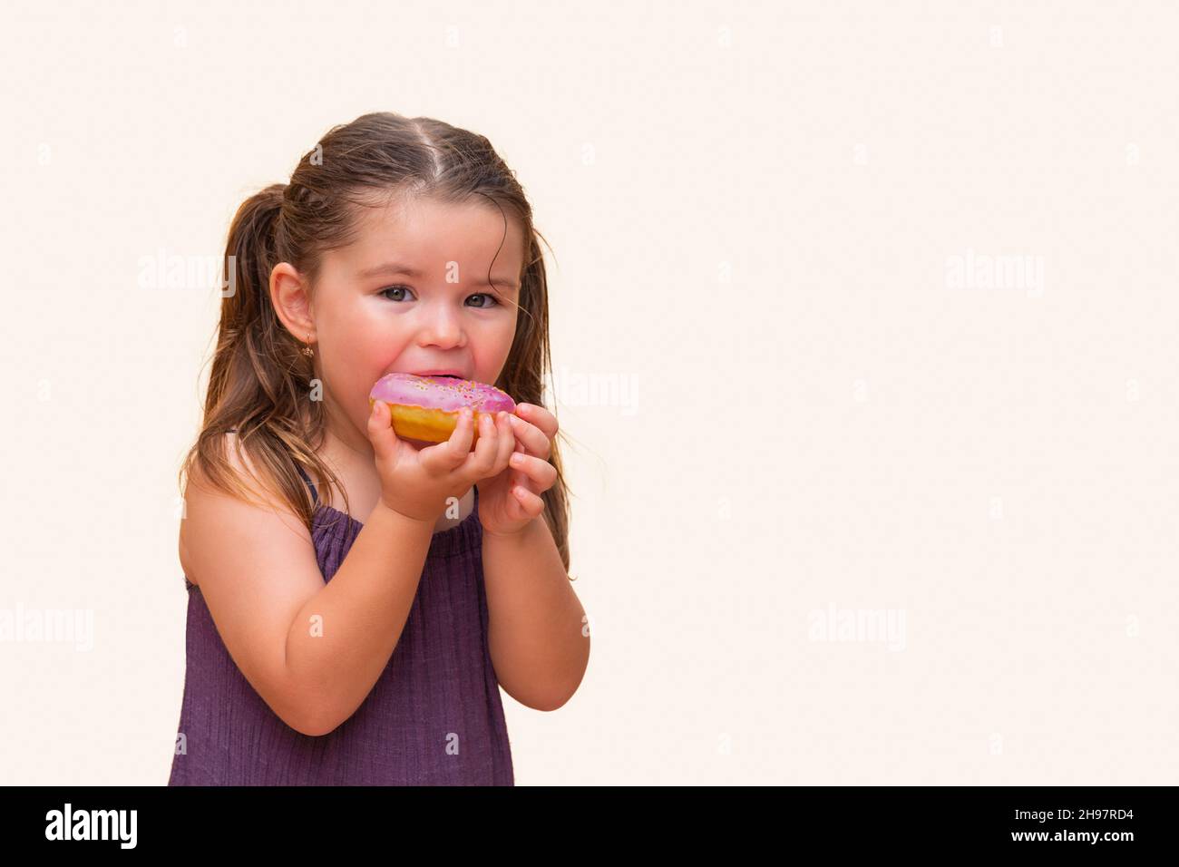 cute little girl in a dress bites a delicious, sweet, pink donut.  child smiles in anticipation of a harmful, tasty meal on a yellow orange background Stock Photo