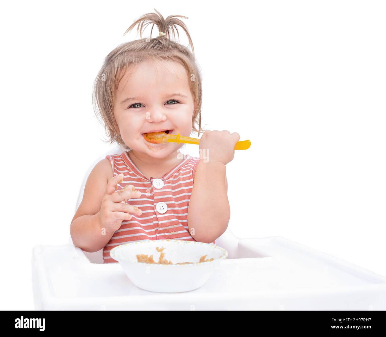 Baby sitting in chair and eating porridge with spoon isolated on white background Stock Photo
