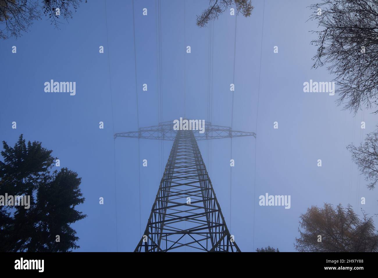 Single Electricity pylon (Strommasten) also overhead line pylon in the forest. Foggy Winter morning and sunset. Up view. Stock Photo