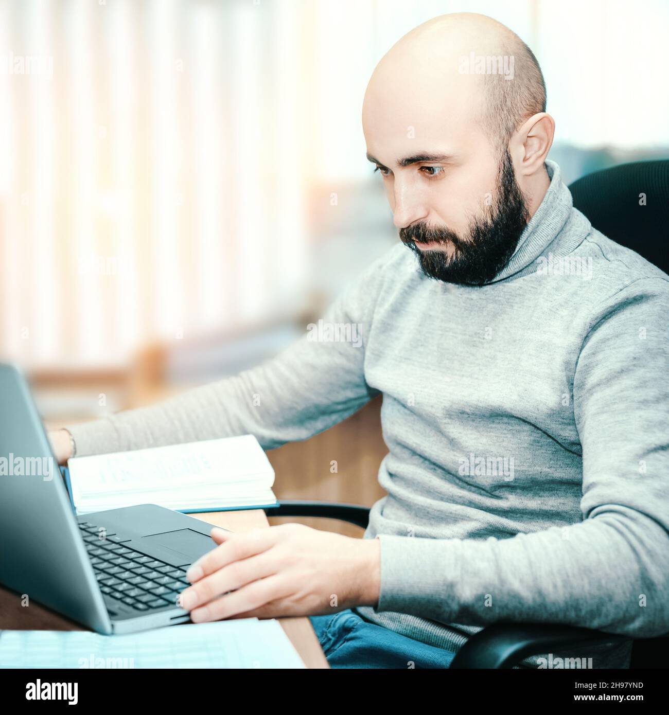 Young bald and bearded man of 25-35 years old works in office for laptop. Guy in casual clothes looks at laptop monitor. Real scene. Real worker. Stock Photo
