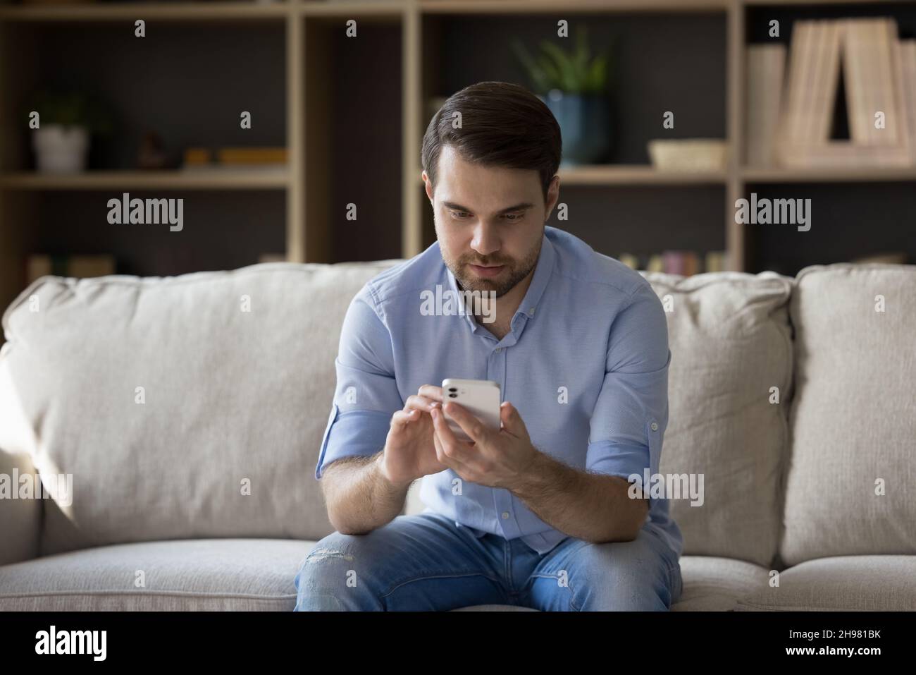 Focused millennial guy un casual using cellphone on couch Stock Photo