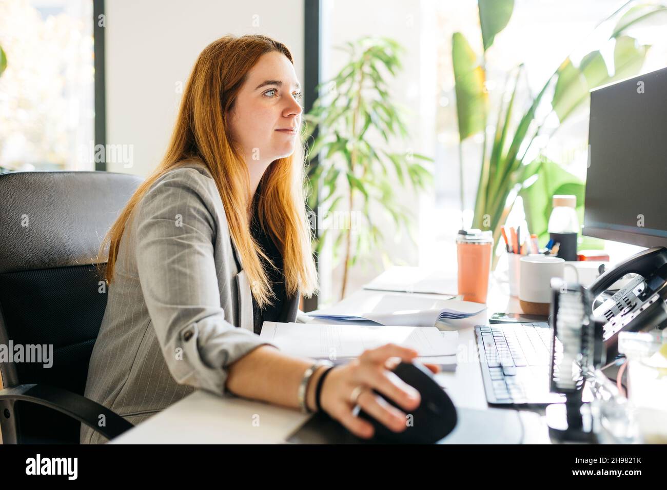 Young red haired woman, sitting on a desk and using a computer, in modern office Stock Photo