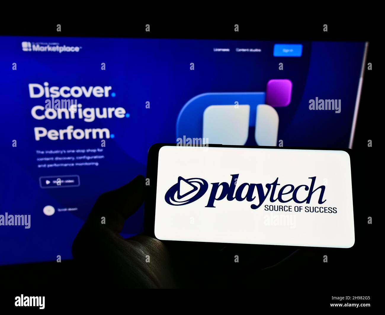 Person holding mobile phone with logo of British gambling software company Playtech plc on screen in front of web page. Focus on phone display. Stock Photo