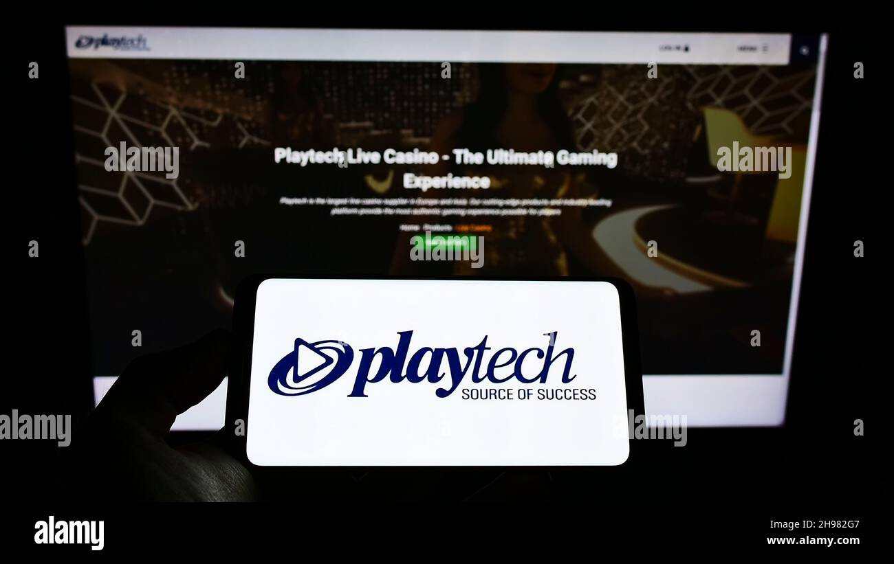 Person holding cellphone with logo of British gambling software company Playtech plc on screen in front of business webpage. Focus on phone display. Stock Photo