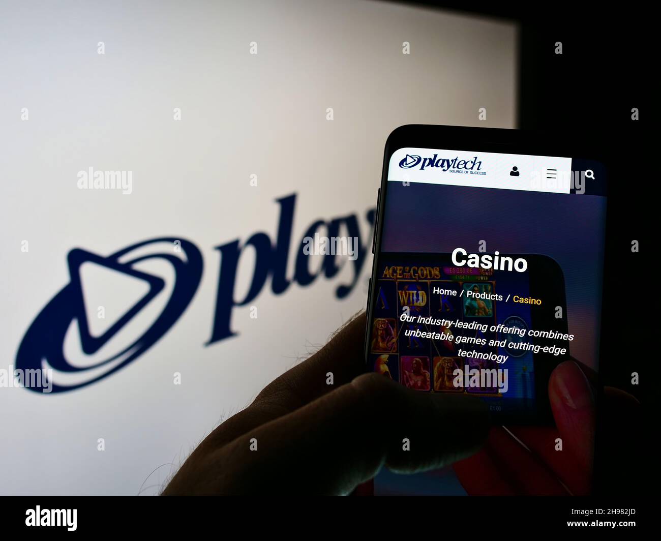 Person holding cellphone with webpage of British gambling software company Playtech plc on screen in front of logo. Focus on center of phone display. Stock Photo