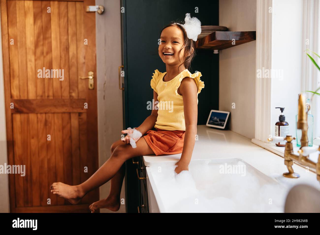 Happy little girl playing with soap bubbles in the kitchen. Cute little girl having fun with white foam while sitting next to the kitchen sink at home Stock Photo