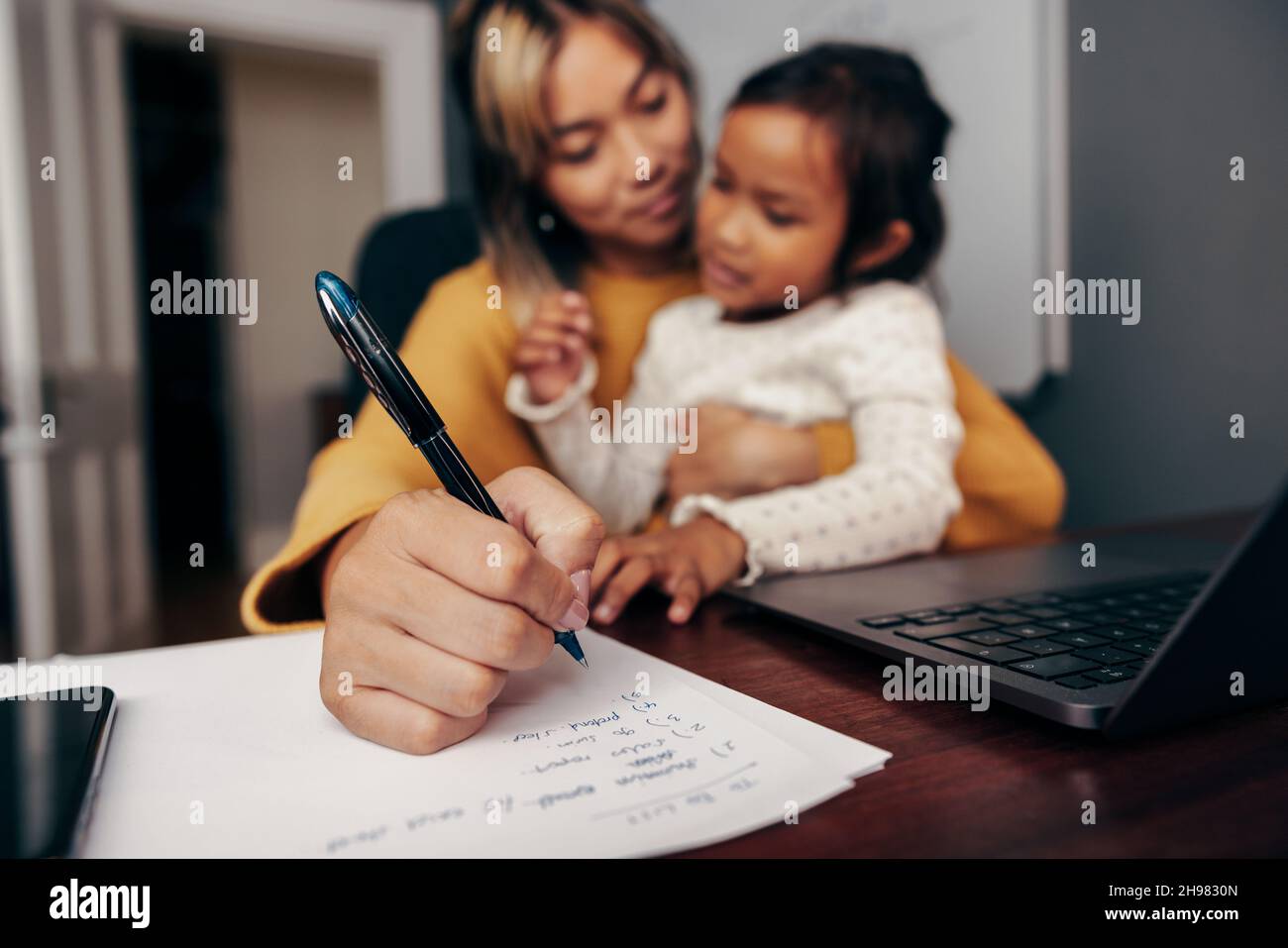 Working mom writing notes while carrying her daughter on her lap. Multi-tasking mom making business plans while sitting at her desk in her home office Stock Photo