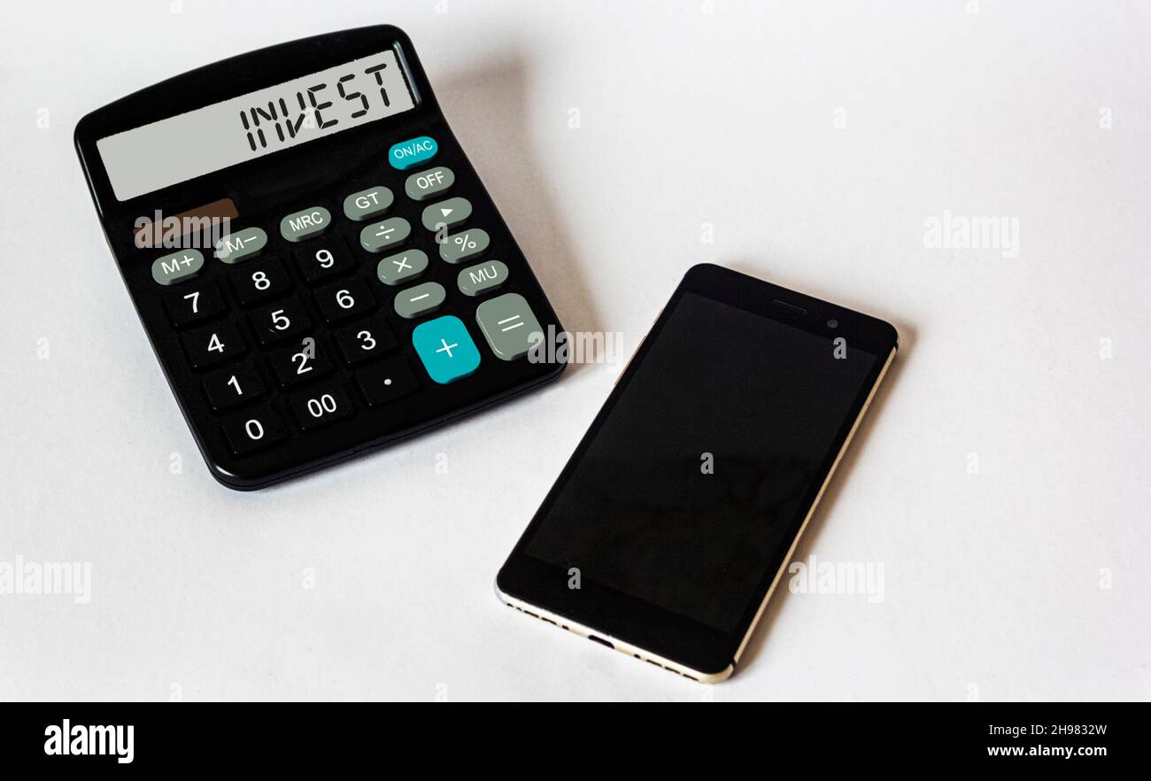 Word invest on calculator display on white background with mobile phone. Financial investments and profit analysis or calculation concept. Stock Photo