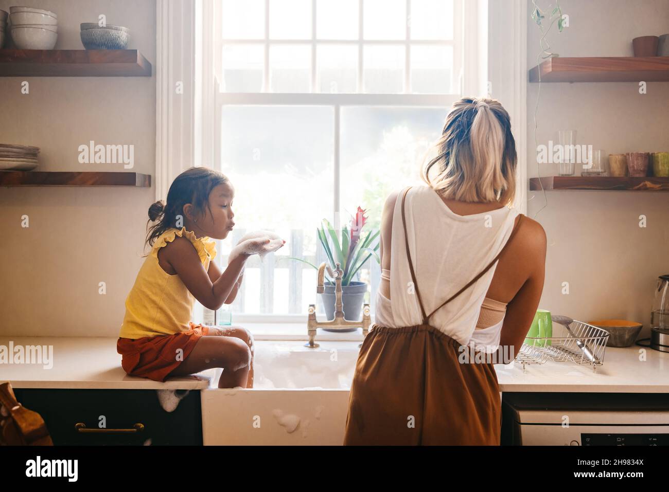 Young girl blowing soap bubbles at her mom in the kitchen. Sweet little girl playing with her mom while she washes the dishes in the sink. Mother and Stock Photo