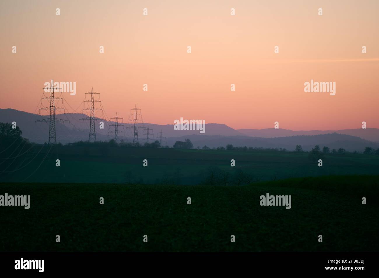 Many Electricity pylons (Strommasten) also overhead line pylon on the field. Green hill landscape. sunset. copy space. Stock Photo