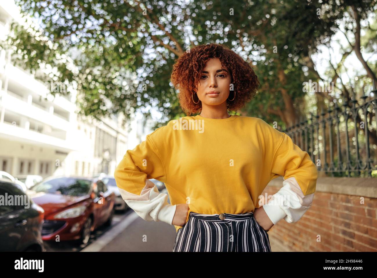 Assertive teenage girl standing on an urban sidewalk with her hands on her hips. Female youngster wearing red hair and a mustard sweatshirt in the cit Stock Photo
