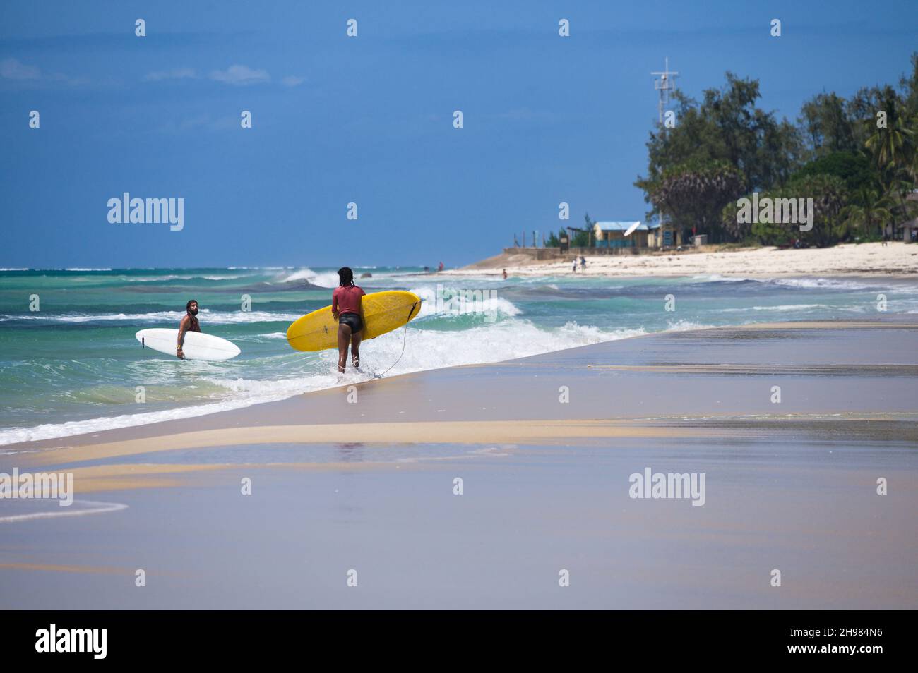 Two surfers walking out of the ocean as waves break on the beach, Diani, Kenya Stock Photo