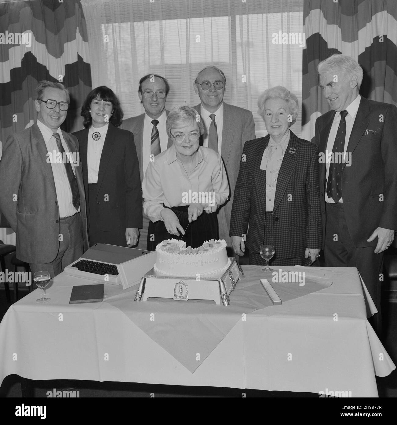John Laing and Son Limited, Page Street, Mill Hill, Barnet, London, 30/03/1987. Mrs Joan Kirby, with a group of friends and colleagues, posed in the middle of cutting the cake during her retirement presentation at Mill Hill. Joan Kirby joined Laing in 1955 as a secretary for Mr Waldrum, who was in charge of personnel. She went on to work for various other individuals including John Renshaw. Joan joined Laing's Welfare Department in 1982 and retired from the Company in 1987, after 32 years of service. The typewriter beside the cake in this image was presented to Joan as a retirement gift. Stock Photo
