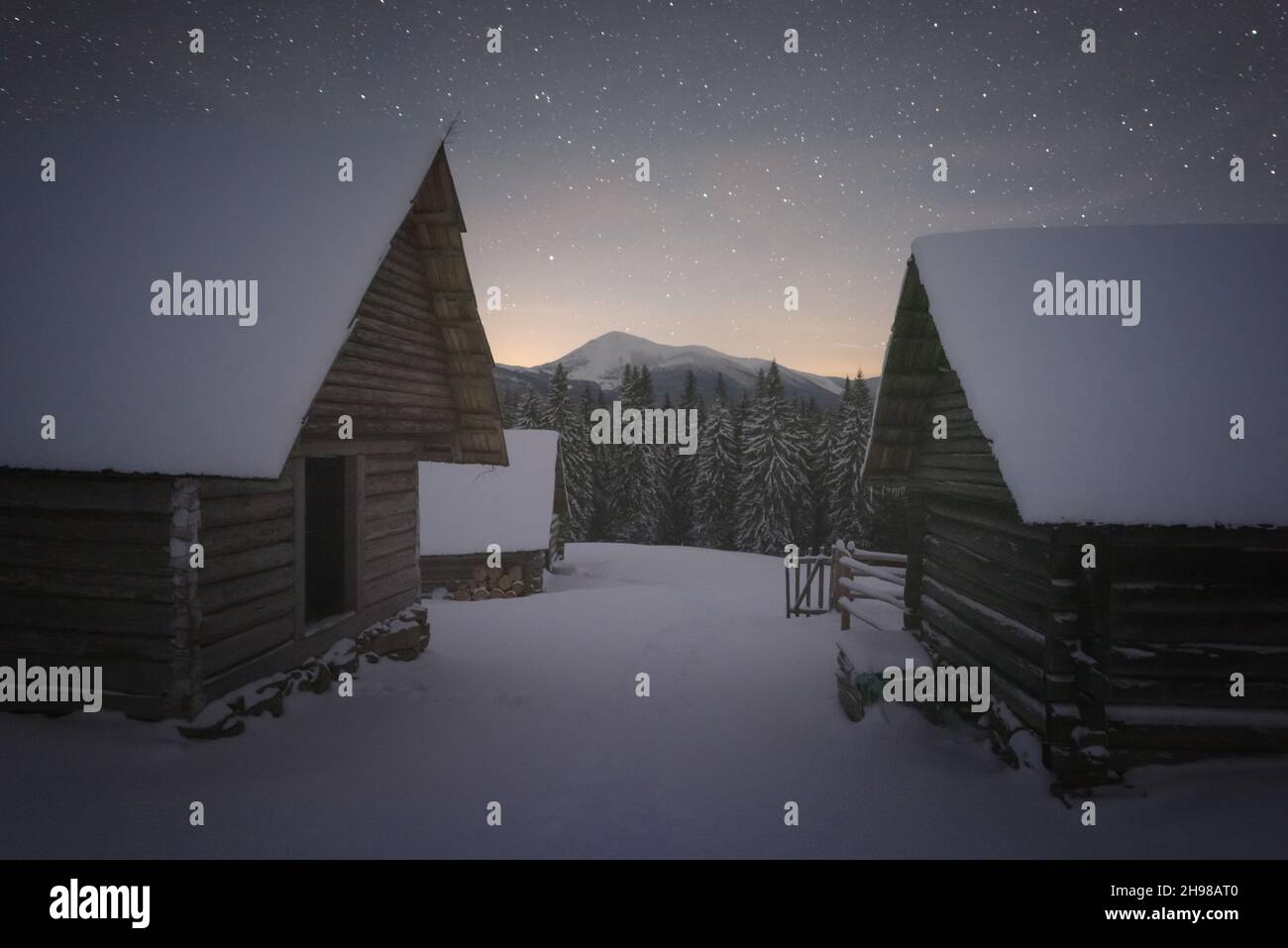 Moody winter landscape with wooden houses in snowy mountains. Starry sky with snowy peaks and snow covered hut. Christmas holiday and winter vacations concept Stock Photo