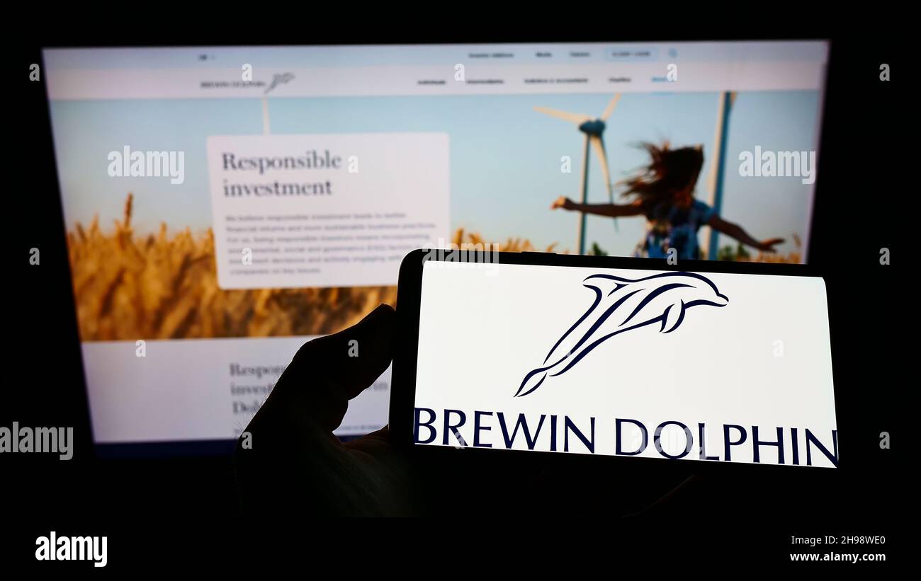 Person holding smartphone with logo of British wealth management company Brewin Dolphin plc on screen in front of website. Focus on phone display. Stock Photo