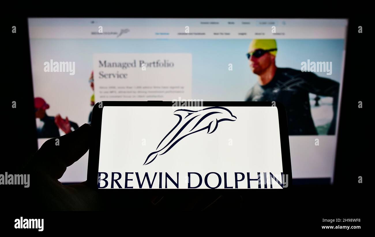 Person holding mobile phone with logo of British wealth management company Brewin Dolphin plc on screen in front of webpage. Focus on phone display. Stock Photo
