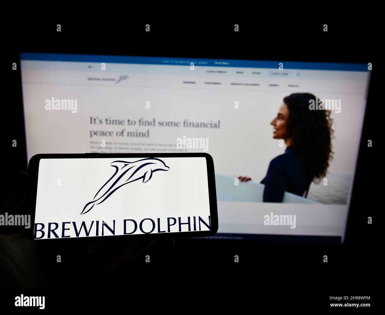 Person holding cellphone with logo of British wealth management company Brewin Dolphin plc on screen in front of web page. Focus on phone display. Stock Photo