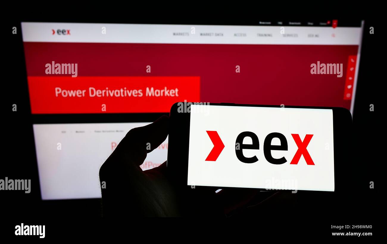 Person holding mobile phone with logo of German company European Energy Exchange AG (EEX) on screen in front of web page. Focus on phone display. Stock Photo