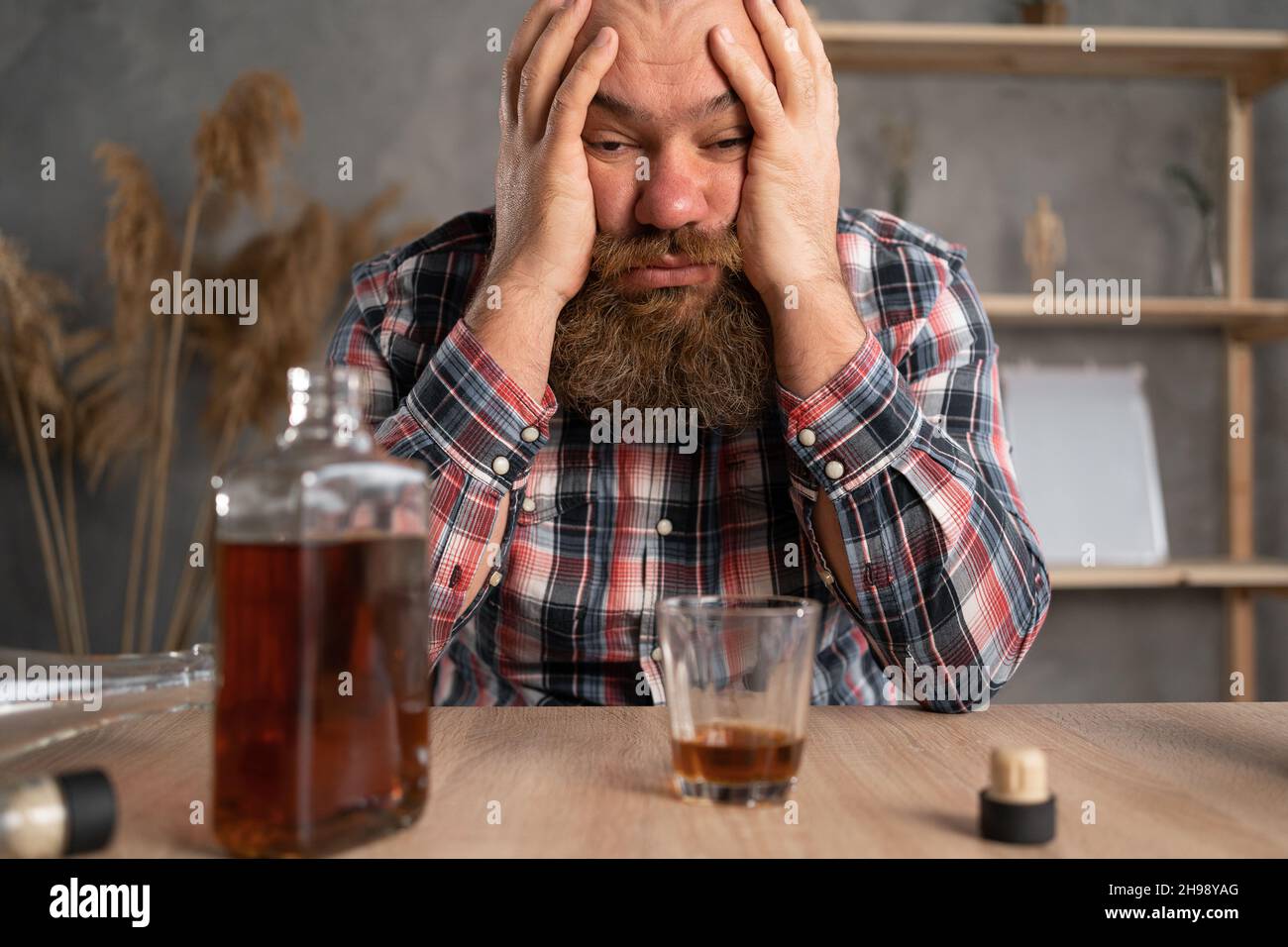 Alcoholism concept. The young man drinks too much alcohol. Sits at home at the table in casual clothes, bowed his head over bottles of cognac and Stock Photo