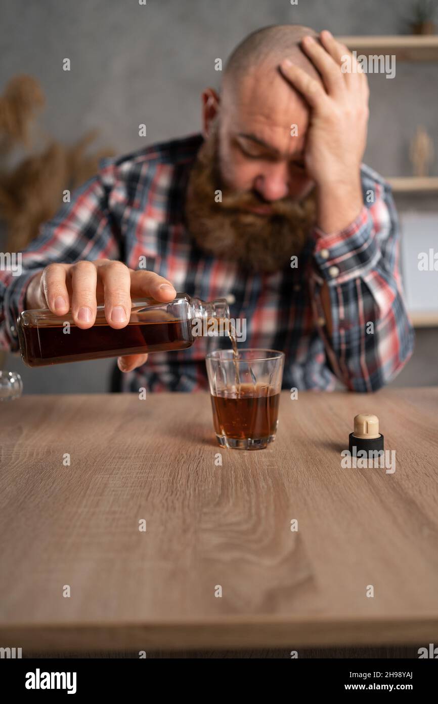 Depression and alcoholism in men. An adult bearded guy himself sits at a table with bottles of alcohol. He holds brandy in his hands and pours it into Stock Photo