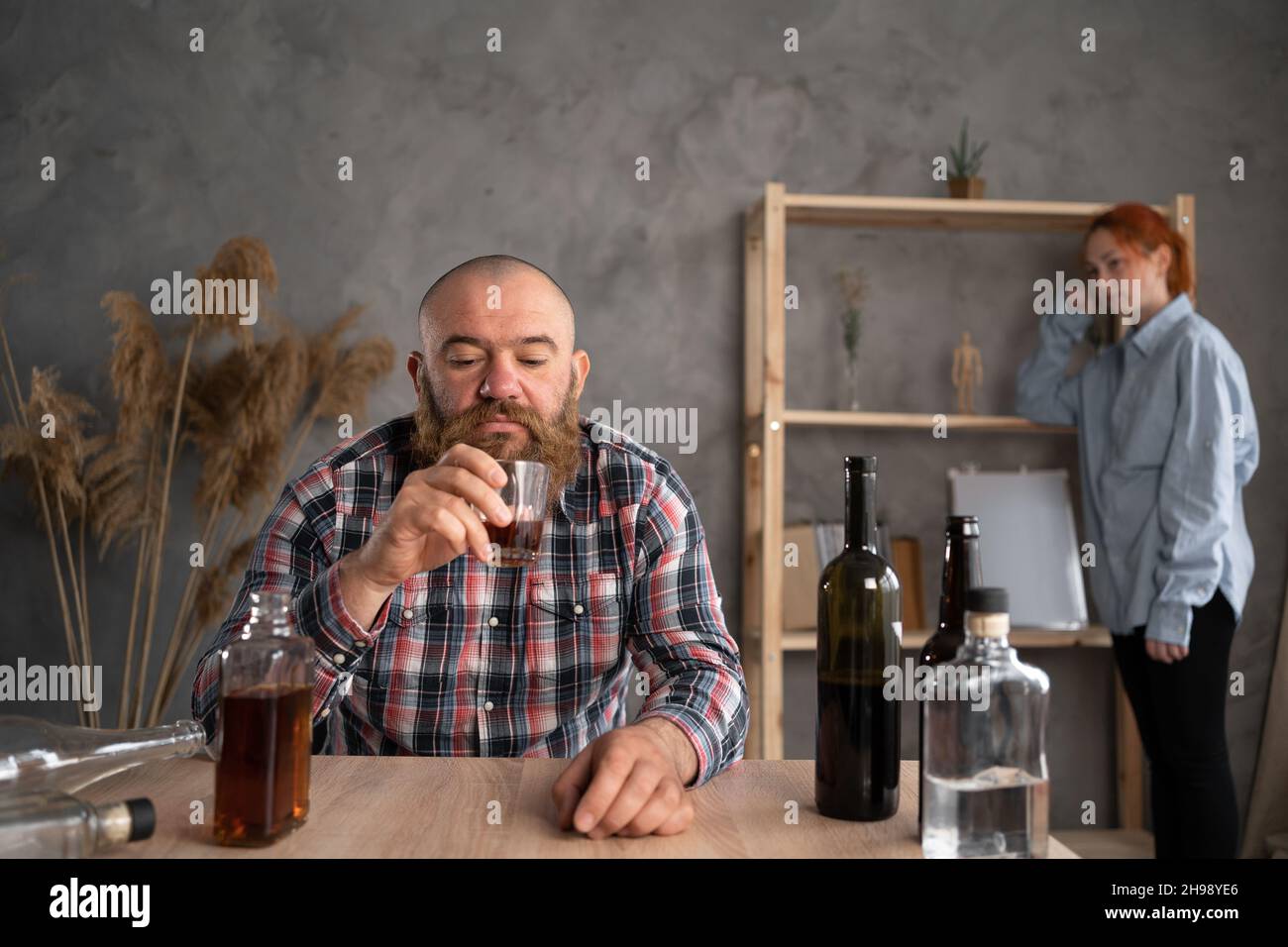 alcoholism in the family, a sad drunk husband pours alcohol into a glass and a sad relaxed wife in the background, alcohol abuse at home. Addiction Stock Photo