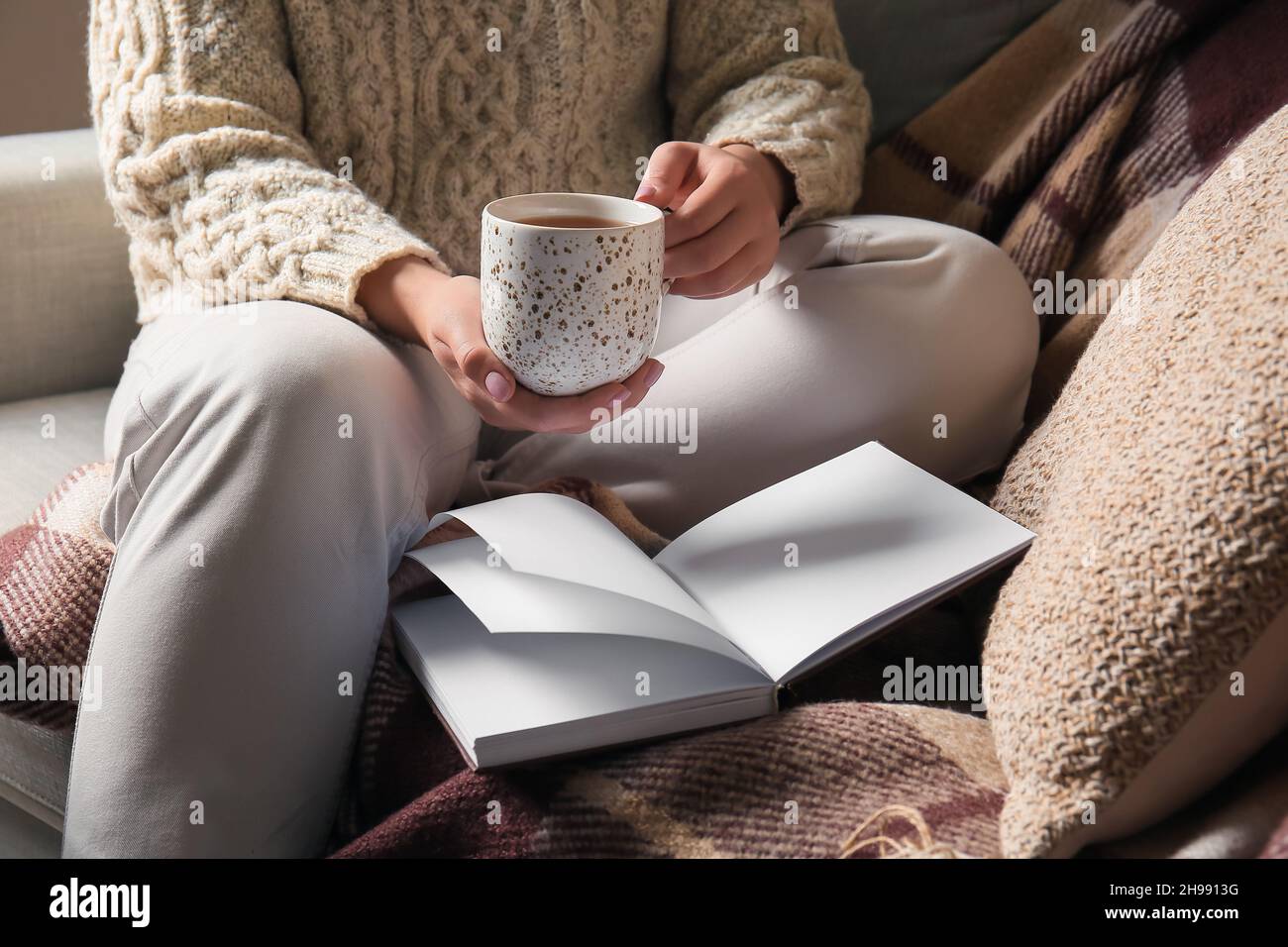 Woman sitting on sofa with opened book and holding cup of tea in living ...