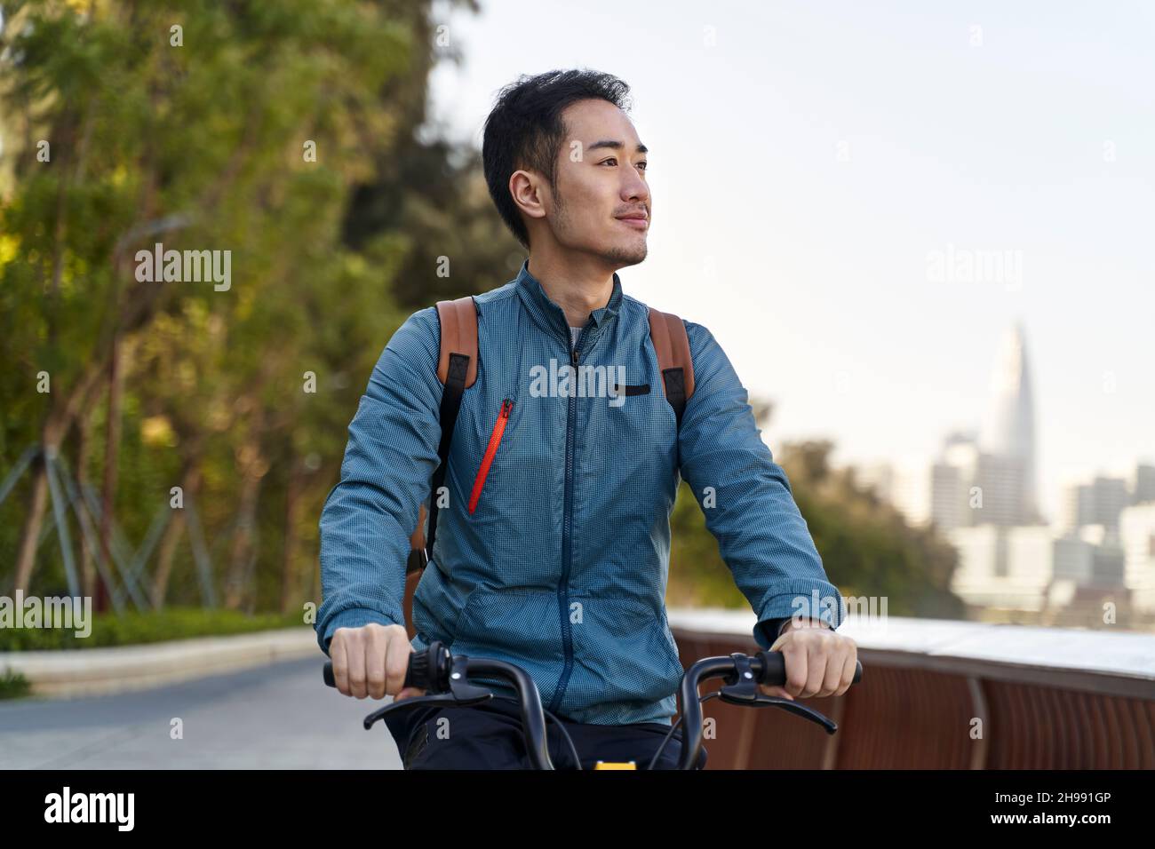young asian man riding bike on riverfront path in public park in modern city Stock Photo
