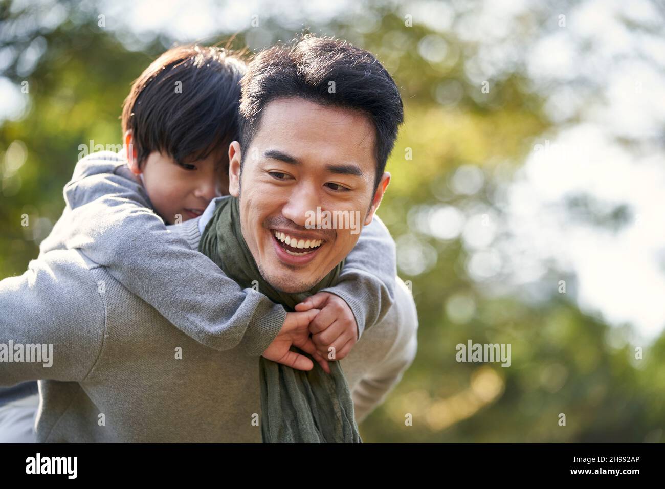 asian father carrying son on back having fun outdoors in park Stock Photo