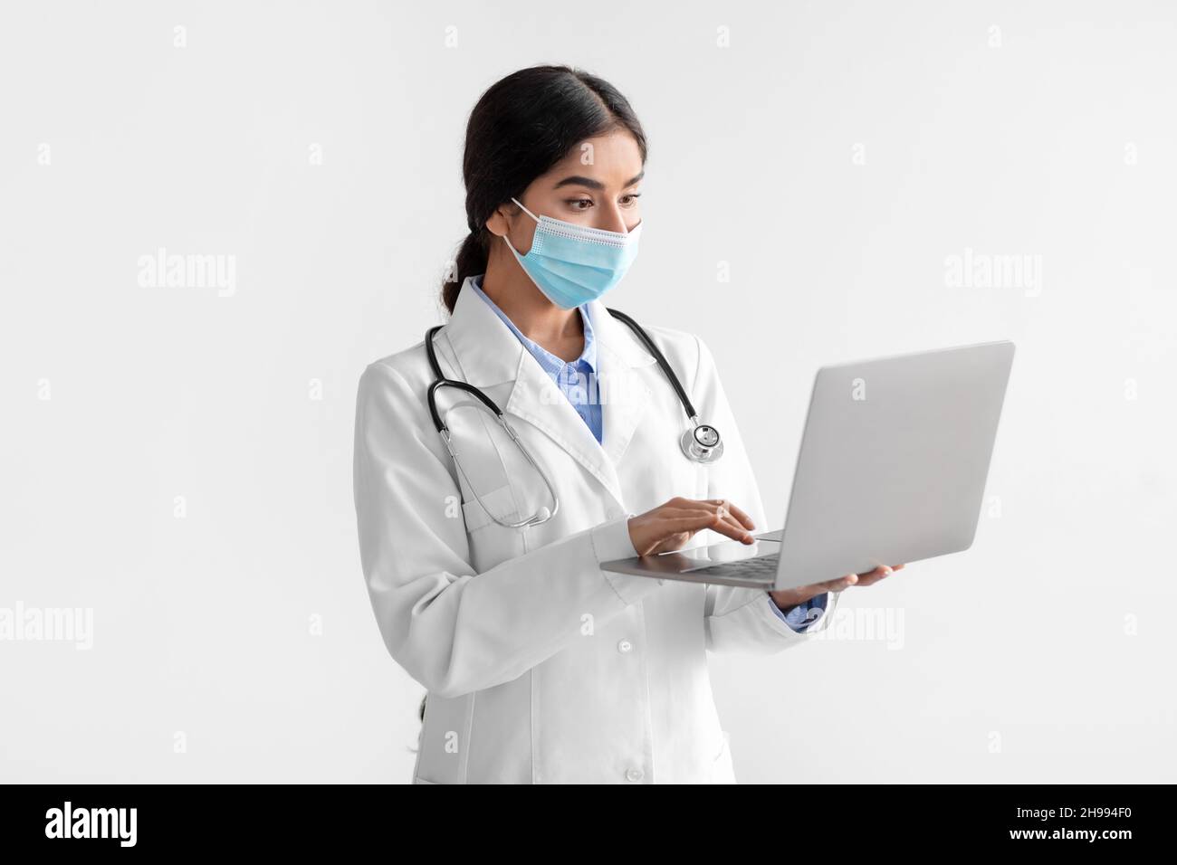 Busy young indian lady doctor in white coat and protective mask working ...