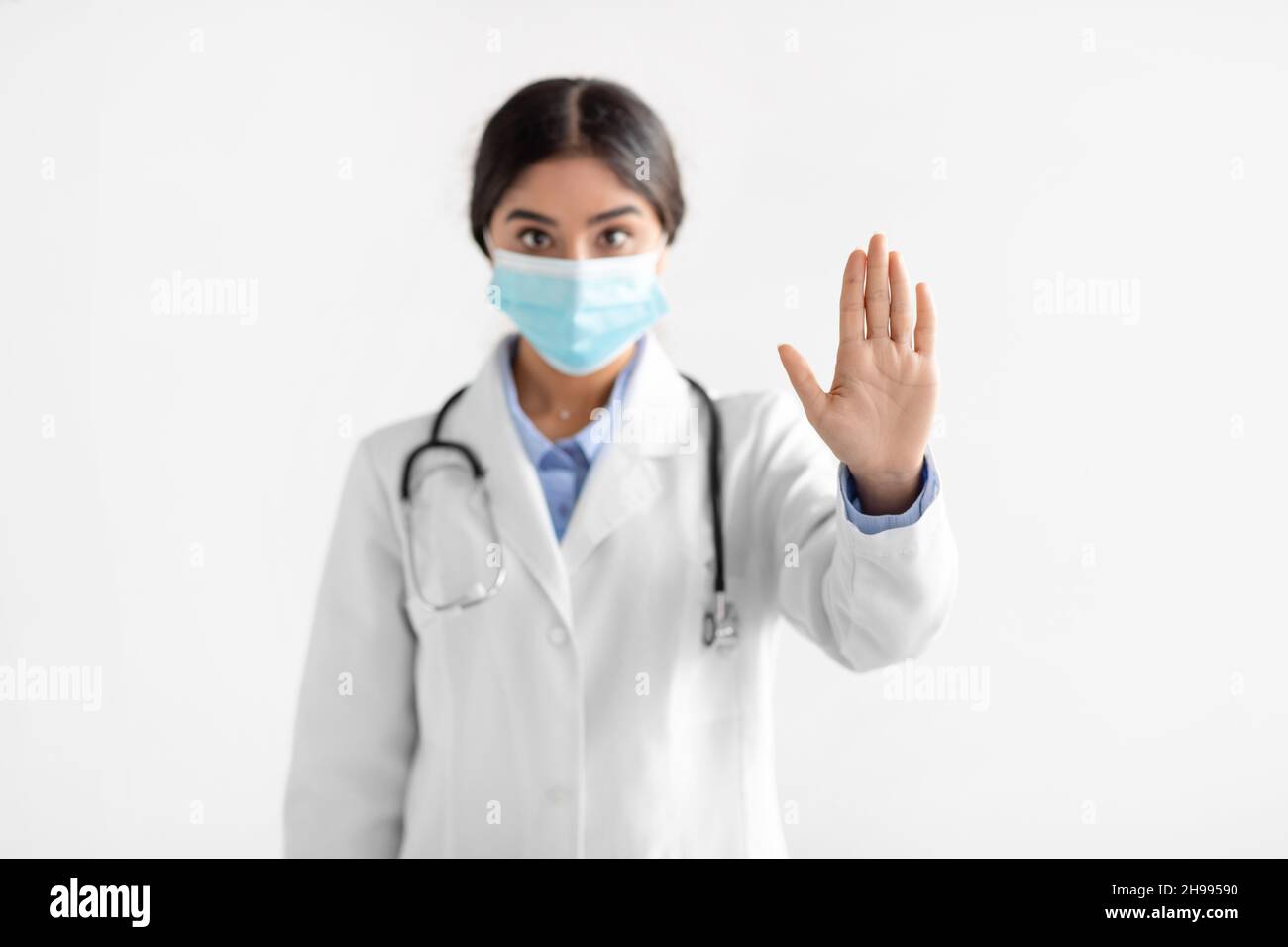 Serious young indian woman doctor in coat and protective mask show stop ...