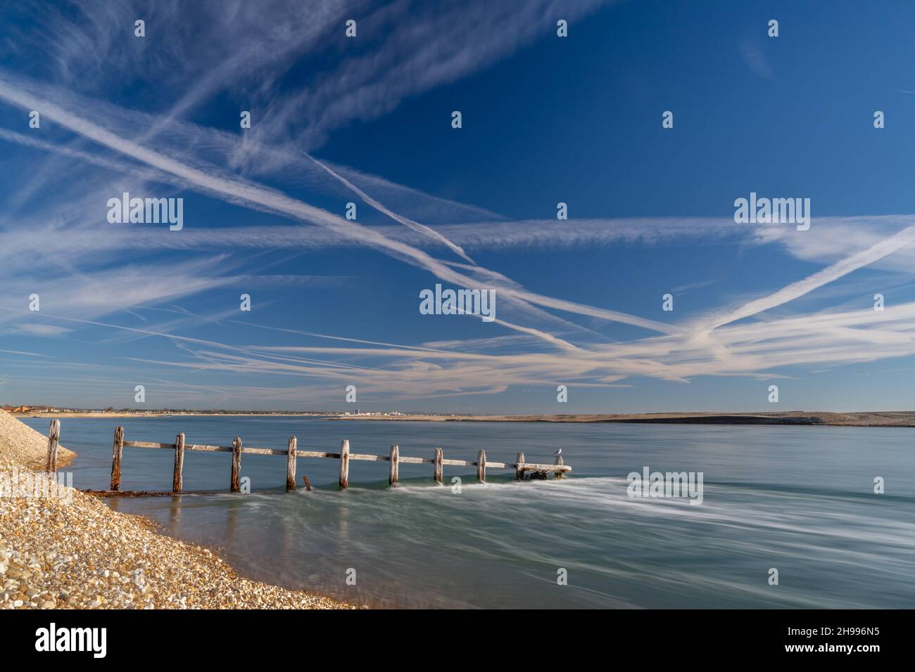 Water rushing into the entrance of Pagham harbour with an old groin looking towards Bognor Regis on a blue sky day with con trails. Stock Photo