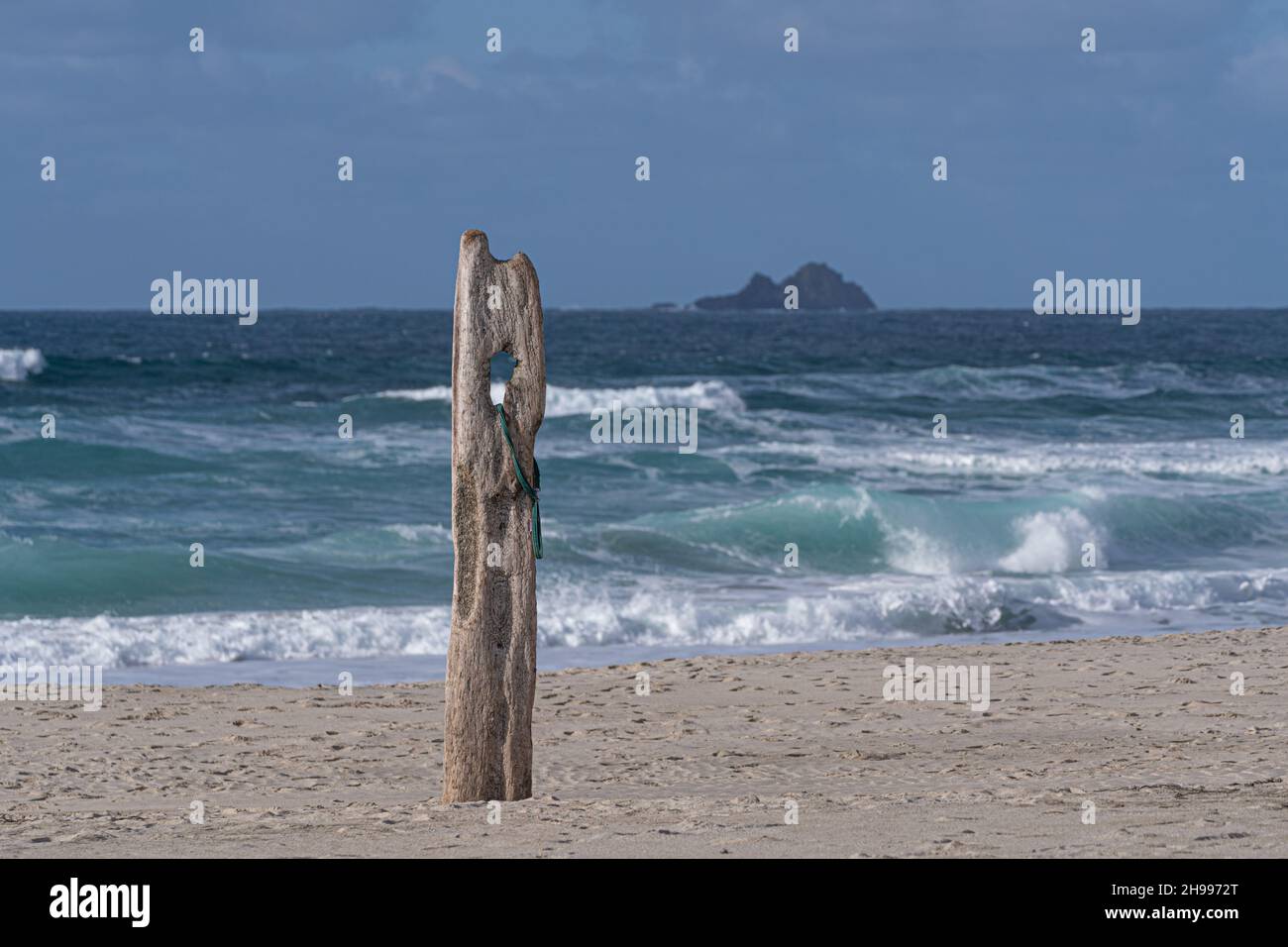 An old decayed grey piece of wood sticking out of the sand on Sennen Cove Beach in Cornwall with the Brison Islands in the distance. Stock Photo