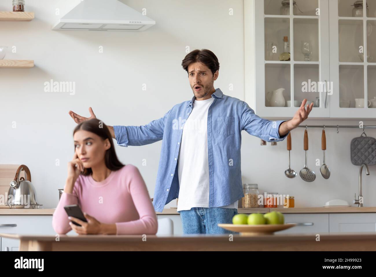 Sad young european woman with smartphone ignores offended angry screaming gesticulating husband Stock Photo