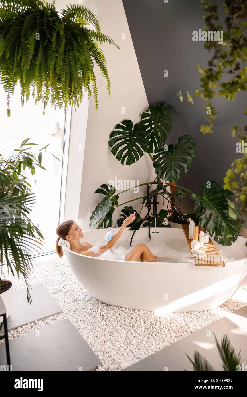 Happy young woman using loofah sponge with applied shower gel while bathing in modern bathroom decorated with tropical plants Stock Photo