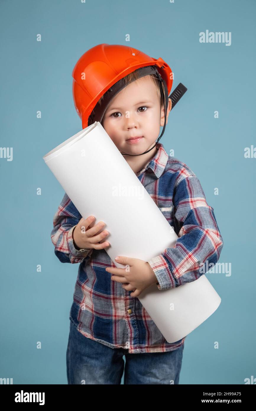 Portrait of adorable little caucasian boy builder in hardhats holding construction drawing against blue background, showing thumb up. The concept of c Stock Photo